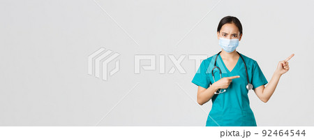 Covid-19, coronavirus disease, healthcare workers concept. Frowning suspicious or frustrated asian female doctor, wear medical mask and scrubs, pointing upper right corner, white background Covid-19, coronavirus disease, healthcare workers concept. Frowning suspicious or frustrated asian female doctor, wear medical mask and scrubs, pointing upper right corner, white background 92464544