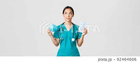 Covid-19, coronavirus disease, healthcare workers concept. Thoughtful smiling asian female doctor, physician in scrubs looking dreamy upper left corner, holding respirator and medical mask Covid-19, coronavirus disease, healthcare workers concept. Thoughtful smiling asian female doctor, physician in scrubs looking dreamy upper left corner, holding respirator and medical mask 92464606
