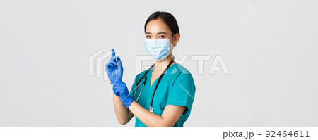 Covid-19, coronavirus disease, healthcare workers concept. Professional confident asian female doctor, nurse in medical mask and scrubs, put on rubber gloves for examination, white background 92464611