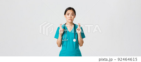 Covid-19, healthcare workers, pandemic concept. Hopeful nervous asian female intern in scrubs waiting for news, doctor cross fingers and biting lip as looking at camera, praying 92464615