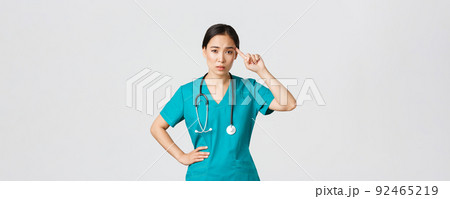Covid-19, healthcare workers, pandemic concept. Annoyed skeptical asian female nurse or doctor scolding someone acting crazy or stupid, roll finger over temple with judgemental face 92465219