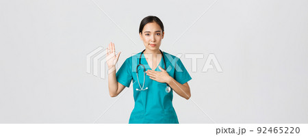 Covid-19, healthcare workers and preventing virus concept. Smiling honest asian female doctor, intern in scrubs giving pledge, raising one arm and hold hand on heart while promise, oath to patient 92465220