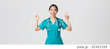 Covid-19, healthcare workers, pandemic concept. Amused cheerful asian female nurse, doctor or surgeon in scrubs pointing fingers and looking up with satisfied face, standing white background 92465399