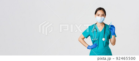 Covid-19, coronavirus disease, healthcare workers concept. Smiling confident asian female nurse in scrubs and medical mask, holding syringe with vaccine, doing vaccination shot, white background 92465500
