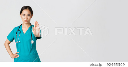 Covid-19, healthcare workers, pandemic concept. Angry serious-looking asian doctor, female physician or nurse in scrubs frowning displeased, extend hand to show stop, disagree, prohibit or forbid 92465509
