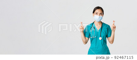 Covid-19, coronavirus disease, healthcare workers concept. Hopeful serious-looking asian doctor, female physician in medical mask and scrubs cross fingers, looking upper left corner 92467115