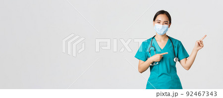 Covid-19, coronavirus disease, healthcare workers concept. Pleasant asian female doctor, medical employee in scrubs and mask, pointing fingers upper right corner, showing advertisement 92467343