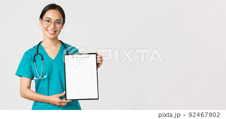 Covid-19, coronavirus disease, healthcare workers concept. Smiling pretty asian female physician, nurse showing test results or contract on clipboard, standing white background 92467902