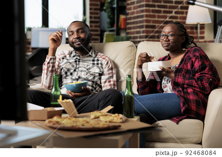 Man and woman enjoying fast food delivery with movie on tv, having fun with leisure activity. Eating takeaway meal with noodles, pizza and chips, drinking beer from bottle. Watching television. 92468084