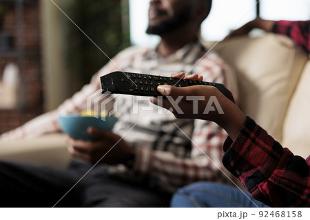 Young woman using tv remote control to switch channel program and find movie on television to watch. Eating takeaway delivery from fast food and watching film together. Close up. 92468158