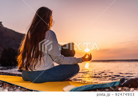 silhoute of Young woman with long hair in blue jeans and sportswear practicing outdoors on yoga mat by the sea on a sunset. Women's yoga fitness routine. Healthy lifestyle, harmony and meditation 92474933