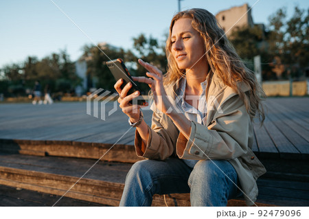 Young casual woman in coat sitting on bench on sea embankment using smartphone 92479096