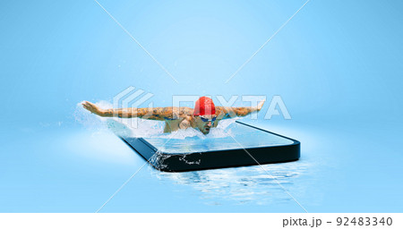 Creative collage with strong swimmer in goggles swimming by butterfly style in water in 3d phone screen over blue background. Sport, competition, online watching sports event Creative collage with strong swimmer in goggles swimming by butterfly style in water in 3d phone screen over blue background. Sport, competition, online watching sports event 92483340