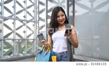 Smiling young woman with shopping bags, using smartphone and walking outside of shopping mall 92486139