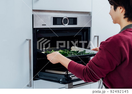 Woman putting baking sheet with teared curly green kale leaves into oven the modern kitchen. Cooking kale chips. Healthy eating, dieting. Step by step cooking. Selective focus, copy space 92492145