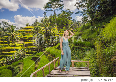 Beautiful young woman walk at typical Asian hillside with rice farming, mountain shape green cascade rice field terraces paddies. Ubud, Bali, Indonesia. Bali travel concept Beautiful young woman walk at typical Asian hillside with rice farming, mountain shape green cascade rice field terraces paddies. Ubud, Bali, Indonesia. Bali travel concept 92496347