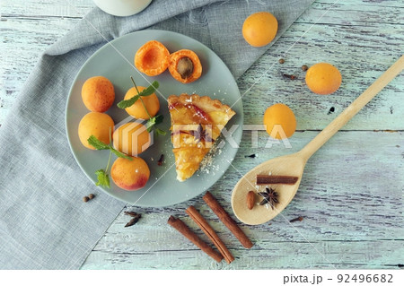 A piece of apricot pie on a plate, sprinkled with powdered sugar, spices and mint leaves, fresh apricots on the kitchen table, homemade food, sweet dessert A piece of apricot pie on a plate, sprinkled with powdered sugar, spices and mint leaves, fresh apricots on the kitchen table, homemade food, sweet dessert 92496682