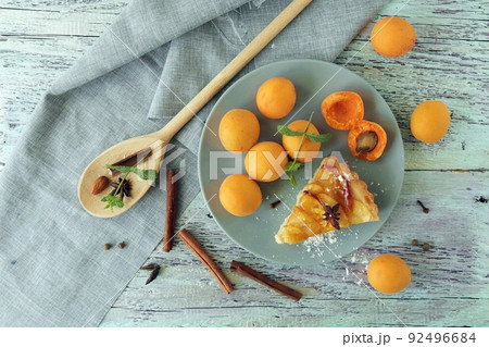 A piece of apricot pie on a plate, sprinkled with powdered sugar, spices and mint leaves, fresh apricots on the kitchen table, homemade food, sweet dessert A piece of apricot pie on a plate, sprinkled with powdered sugar, spices and mint leaves, fresh apricots on the kitchen table, homemade food, sweet dessert 92496684
