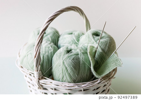Light green yarn threads in a white basket on a gray background with knitting needles. Light green yarn threads in a white basket on a gray background with knitting needles. 92497389