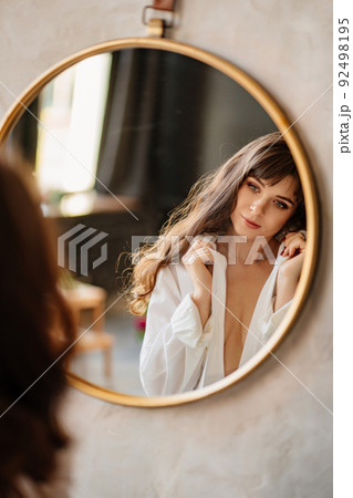 a beautiful woman in a white shirt looks at her reflection in a round mirror. a beautiful woman in a white shirt looks at her reflection in a round mirror. 92498195