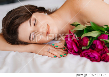 a gentle brunette woman in bed with a bouquet of peonies. morning of holiday. a gentle brunette woman in bed with a bouquet of peonies. morning of holiday. 92498197