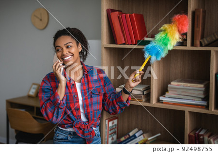 Smiling millennial african american housewife wiping dust, calling by phone in room interior Smiling millennial african american housewife wiping dust, calling by phone in room interior 92498755
