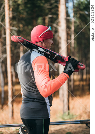 Training an athlete on the roller skaters. Biathlon ride on the roller skis with ski poles, in the helmet. Autumn workout. Roller sport. The athlete goes and holds sports equipment in his hand. 92511804