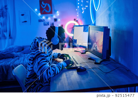 African gamer boy in wireless headphones typing on keyboard while sitting at table in front of computer and playing video game 92515477