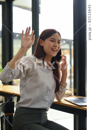 Cheerful female employee waving hands to greet her colleagues while talking on mobile phone 92515645