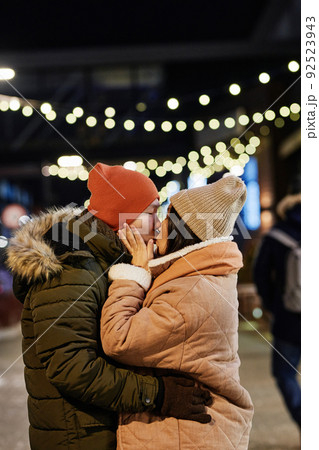 Vertical medium shot of young Asian man and woman in love wearing casual warm clothes standing outdoors on winter evening kissing each other 92523943