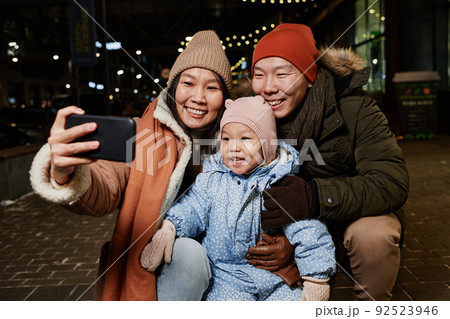 Horizontal shot of Asian woman taking selfie photos with her husband and child on smartphone camera on winter evening 92523946