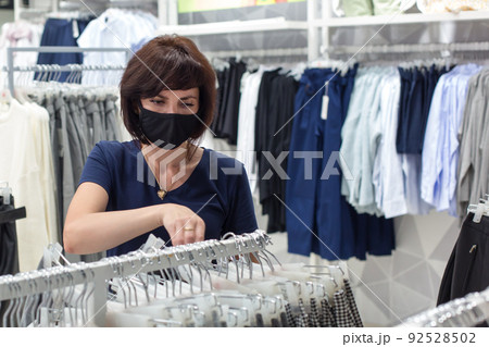 A brunette woman in a black protective mask in the store chooses her clothing size A brunette woman in a black protective mask in the store chooses her clothing size 92528502