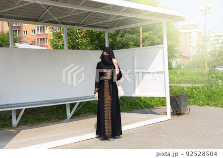 A Muslim woman in black national clothes stands alone at a bus stop in the city A Muslim woman in black national clothes stands alone at a bus stop in the city 92528504