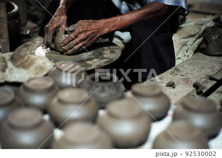 Selective focused on the dirty wrinkled skin hands of old man molding the clay work on the spinning wheel for making the jar with blurred group of clay jars in foreground 92530002