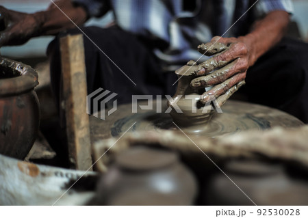 Selective focused on the dirty wrinkled skin hands of old man molding the clay work on the spinning wheel for making the jar with blurred group of clay jars in foreground Selective focused on the dirty wrinkled skin hands of old man molding the clay work on the spinning wheel for making the jar with blurred group of clay jars in foreground 92530028