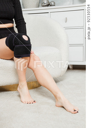 Black knee support brace on a woman leg. girl in an orthosis in the interior of the house. Orthopedic anatomic braces for knee fixation, injuries and pain. Knee Joint Bandage Sleeve. Elastic Sports. Black knee support brace on a woman leg. girl in an orthosis in the interior of the house. Orthopedic anatomic braces for knee fixation, injuries and pain. Knee Joint Bandage Sleeve. Elastic Sports. 92530554