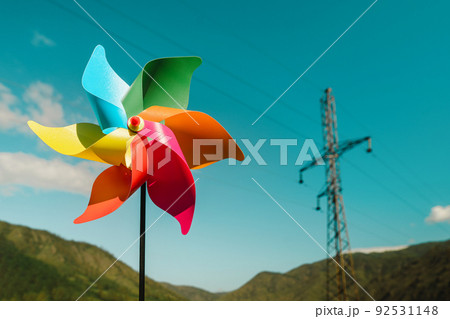 Close-up multi-colored pinwheel and electric power line against the background of green mountains and sky. Close-up multi-colored pinwheel and electric power line against the background of green mountains and sky. 92531148