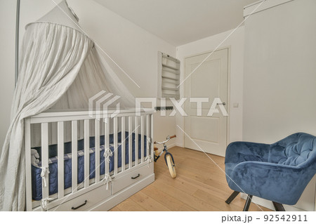 Interior of white room with baby crib and armchair near window with blue chest of drawers in daylight Interior of white room with baby crib and armchair near window with blue chest of drawers in daylight 92542911