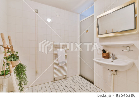 Cupboard with sink and mirror attached to tiled wall near door and drying rack in modern restroom 92545286
