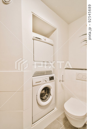 Modern toilet installed on beige wall under button and illuminated shelf with washing machine in light restroom at home 92545689
