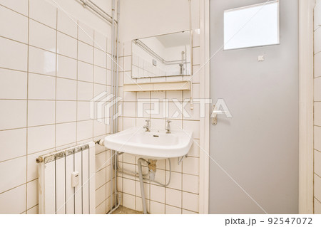 Cupboard with sink and mirror attached to tiled wall near door and drying rack in modern restroom Cupboard with sink and mirror attached to tiled wall near door and drying rack in modern restroom 92547072