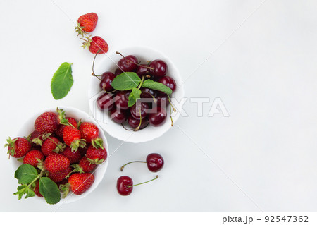 Two white plates with cherries and strawberries with mint petals stand on a white background with space for text. High quality photo Two white plates with cherries and strawberries with mint petals stand on a white background with space for text. High quality photo 92547362