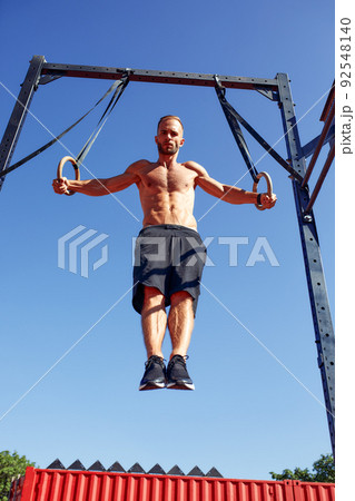 Handsome man on sport ground. The sportsman the guy, carries out difficult exercise, sports gymnastics. Muscular man doing workout with rings. The concept of a modern young man 92548140
