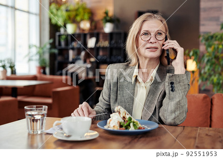 Mature businesswoman in eyeglasses talking to her clients on mobile phone while sitting at the table and eating dessert at the restaurant Mature businesswoman in eyeglasses talking to her clients on mobile phone while sitting at the table and eating dessert at the restaurant 92551953