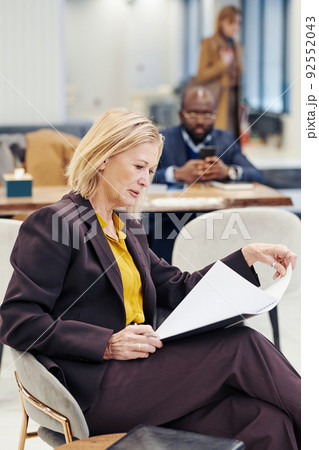 Mature female lawyer in suit sitting on chair and examining the document before meeting with her colleagues in the background 92552043