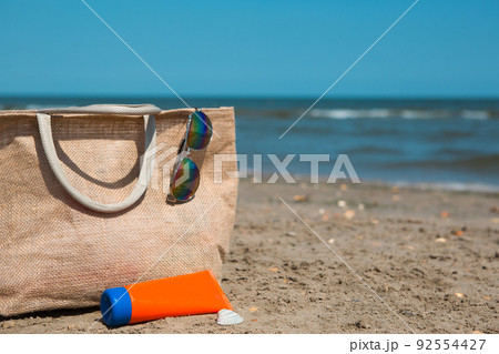 An orange tube with mock up sunscreen, sunglasses and a beach bag stand on the sand near the sea and swing from the breeze. UV protection, beach holidays, travel An orange tube with mock up sunscreen, sunglasses and a beach bag stand on the sand near the sea and swing from the breeze. UV protection, beach holidays, travel 92554427