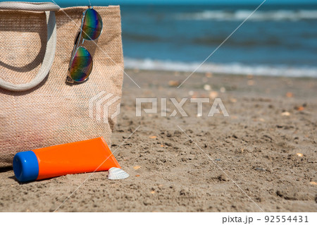 An orange tube with mock up sunscreen, sunglasses and a beach bag stand on the sand near the sea and swing from the breeze. UV protection, beach holidays, travel An orange tube with mock up sunscreen, sunglasses and a beach bag stand on the sand near the sea and swing from the breeze. UV protection, beach holidays, travel 92554431