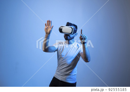 Serious millennial male student in white t-shirt and vr glasses moves his hands manipulating gestures in virtual reality world. 3d technology, virtual reality and entertainment concept. Serious millennial male student in white t-shirt and vr glasses moves his hands manipulating gestures in virtual reality world. 3d technology, virtual reality and entertainment concept. 92555185