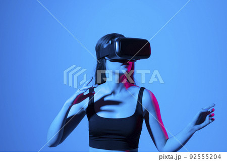 Young brunette woman wearing virtual reality device, vr glasses and playing a game with a friends online in neon light. 92555204