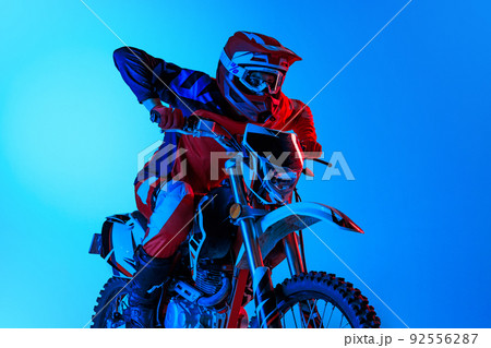 Portrait of young concentrated man, biker riding on cross type motorbike isolated over blue studio background in neon light 92556287